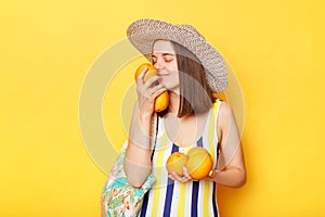 Enjoyable calm woman wearing striped swimsuit and straw hat  yellow background standing holding fresh fruit smelling