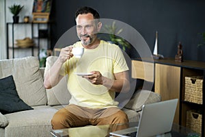 Enjoying coffee or tea middle aged business man make a break while working on laptop sitting on the sofa at home