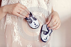 Enjoying future life. Pregnant woman holding small baby booties at her belly, closeup