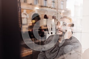 Enjoying his phone call. Handsome young man talking on mobile phone and looking throughwindow with smile while sitting in cafe