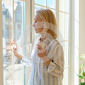 Beautiful senior woman with cup of coffee looking in window while enjoying new day at home