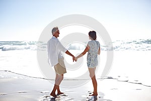 Enjoying the romance of the ocean. Rearview of a happy mature couple walking hand in hand on the beach.
