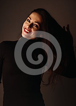 Enjoying toothy smiling makeup woman with brown long hair posing in black sweater on dark shadow background. Closeup