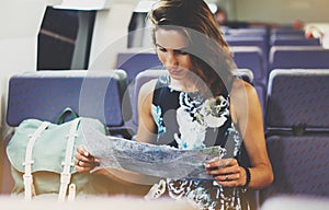 Enjoying travel. Young hipster smile girl with backpack traveling by train sitting near the window holding in hand and looking map