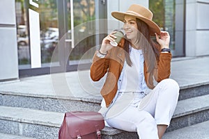 Enjoying vacation. Young and beautiful happy female traveler or tourist wearing hat and backpack drinking coffee and