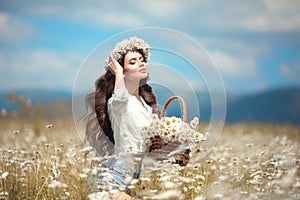 Enjoyment. Beautiful young girl with basket of flowers over chamomile field. Carefree happy brunette woman with healthy wavy hair