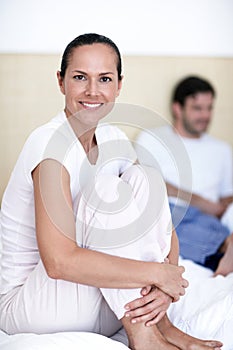 She enjoys the lazy mornings with her husband. A young woman sitting on a bed with her husband in the background.