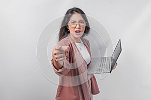 Enraged young Asian business woman holding a laptop pointing to the camera with angry expression, isolated by white background