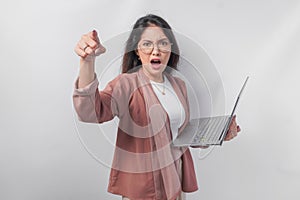 Enraged young Asian business woman holding a laptop pointing to the camera with angry expression, isolated by white background