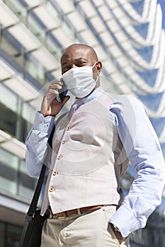 Enterprising black man in elegant clothes and medical mask is talking on the phone next to an office building