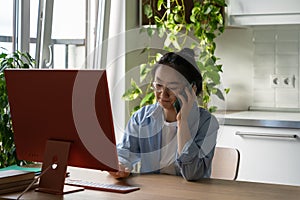 Enterprising pensive Asian woman sits at desk with computer in home office making phone call photo