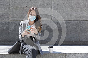 Enterprising young woman with a face mask touches her mobile phone while she is sitting. Next to her she has a coffee. Selective