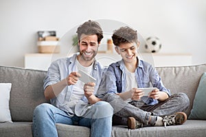 Entertainment and leisure. Father competing with son in online video games on smartphones, sitting together on couch
