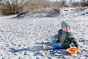 Enthusiast kid sits on a beach playing with sand. Fervid or fervent child dressed against the cold is sitting on a strand