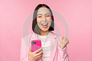 Enthusiastic asian businesswoman saying yes, winning on mobile phone, using smartphone and triumphing, celebrating