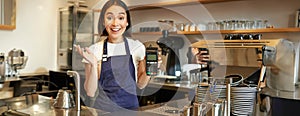 Enthusiastic asian girl barista in apron, showing contactless credit card machine, looks amazed by fast contactless