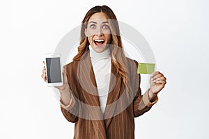 Enthusiastic businesswoman showing her phone screen, app interface and credit card, smiling amazed, white background