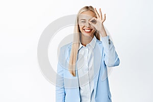 Enthusiastic businesswoman showing zero, okay gesture on eye and smiling, recommending something good, standing in suit