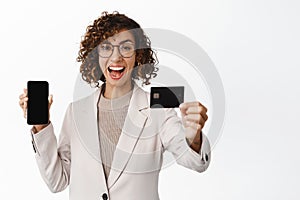 Enthusiastic businesswoman in suit showing mobile phone empty screen, credit card and shopping app interface