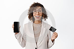 Enthusiastic businesswoman in suit showing mobile phone empty screen, credit card and shopping app interface