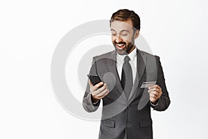 Enthusiastic corporate man looking pleased at his phone, smiling, holding credit card, receive money tranfer in
