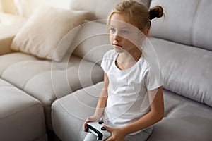 Enthusiastic kid in casual outfit playing with joystick while sitting on sofa in livingroom