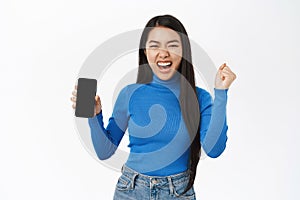 Enthusiastic korean woman shows mobile phone screen, looks excited and surprised, winning on smartphone, standing over