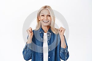 Enthusiastic middle aged woman, senior lady celebrating, triumphing and achieve goal, standing over white background