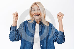 Enthusiastic middle aged woman, senior lady celebrating, triumphing and achieve goal, standing over white background