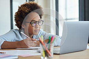 Enthusiastic mixed race teen schoolgirl in glasses listening to her teacher, making notes, using laptop during online