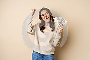 Enthusiastic modern woman winning money, got cash, celebrating and shouting of joy, standing against beige background
