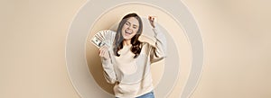 Enthusiastic modern woman winning money, got cash, celebrating and shouting of joy, standing against beige background