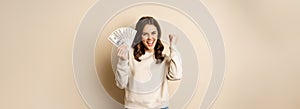 Enthusiastic modern woman winning money, got cash, celebrating and shouting of joy, standing against beige background