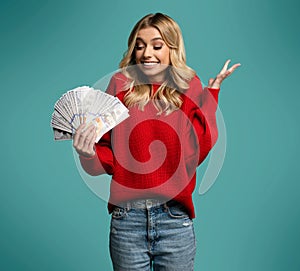 Enthusiastic modern woman winning money, got cash, celebrating and shouting of joy, standing against blue