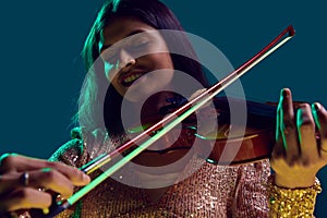 Enthusiastic musician, Beautiful Indian woman in sequined attire playing violin in neon light against gradient