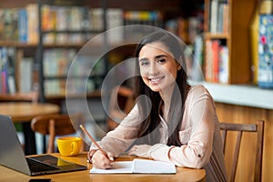 Enthusiastic student latin lady looking for job online, using laptop and taking notes, sitting at cafe, copy space