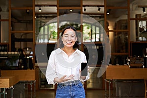 Enthusiastic young asian woman, cafe manager demonstrating app on mobile phone, showing smartphone screen, standing in