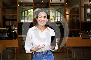 Enthusiastic young asian woman, cafe manager demonstrating app on mobile phone, showing smartphone screen, standing in