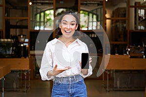 Enthusiastic young asian woman, cafe manager demonstrating app on mobile phone, showing smartphone screen, standing in