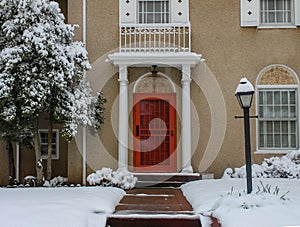 Entrance of elegant upscale stucco house with pillars and tiles in snow with bright red door