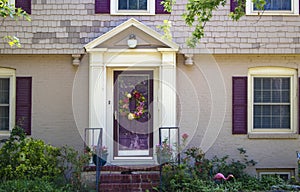 Entrance and front door of beauitufl painted brick and shingle cottage with purple shutters and triming and wreath and roses and p