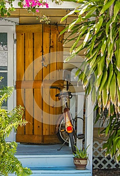 Entrance and porch to Key West House with rustic hurricane shutter by door and rusted bike parked surrounded by tropical greenery