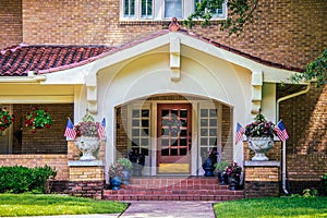 Entrance and stuco bungalow porch with french doors and tile roof on brick house with many flower pots and American flags and star