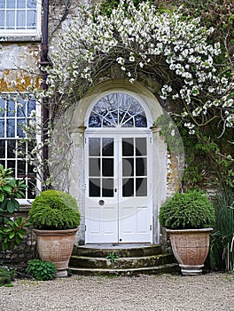 Entrance to a historic manor, framed by antique architectural elements and flanked by potted topiaries, features an aged photo