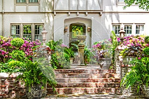 Entrance to up-scale chalet-like stucco house with brick stairs up to flower filled courtyard with fountain and trees reflected in