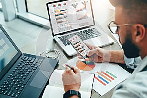 Entrepreneur sits in office,uses smartphone, working on laptop with charts, diagrams on screen, makes notes in notebook