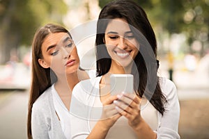 Envious Girl Peeking At Girlfriend`s Phone While She Texting Outdoors