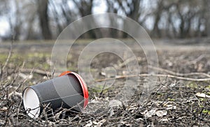 Enviromental pollution; paper cup for coffee and red plastic cover on a background of forest nature and grass.