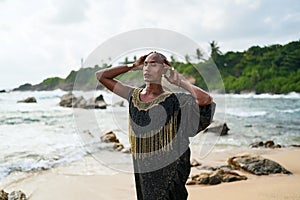 Epatage lgbtq black male posing with hands up on camera on scenic ocean beach. Non-binary ethnic fashion model in long