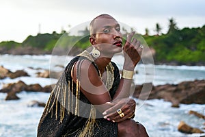 Epatage lgbtq black man poses on scenic ocean beach looks at camera demonstrates jewellery . Androgynous ethnic fashion